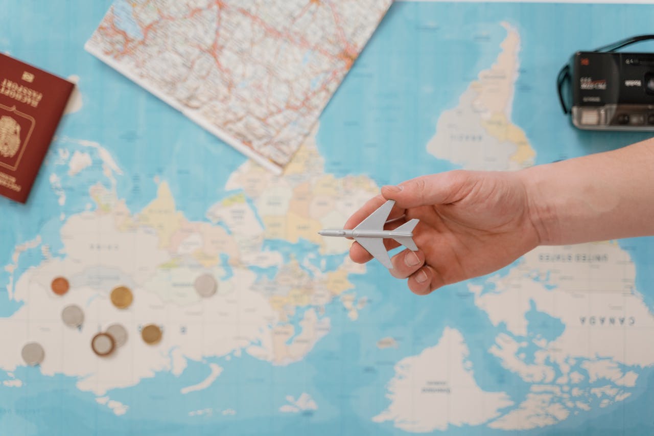 hero-img-02 Hand holding a toy airplane over a world map, surrounded by coins, camera, and passport.